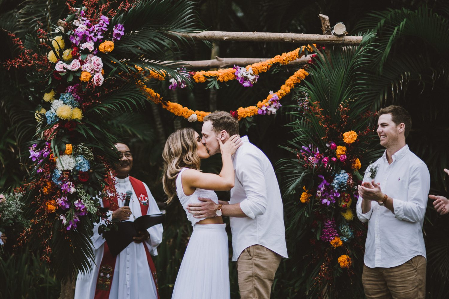 Carly & Nick kissing the ceremony of their wedding Bali, with an exotic backdrop with colorful floral decorations