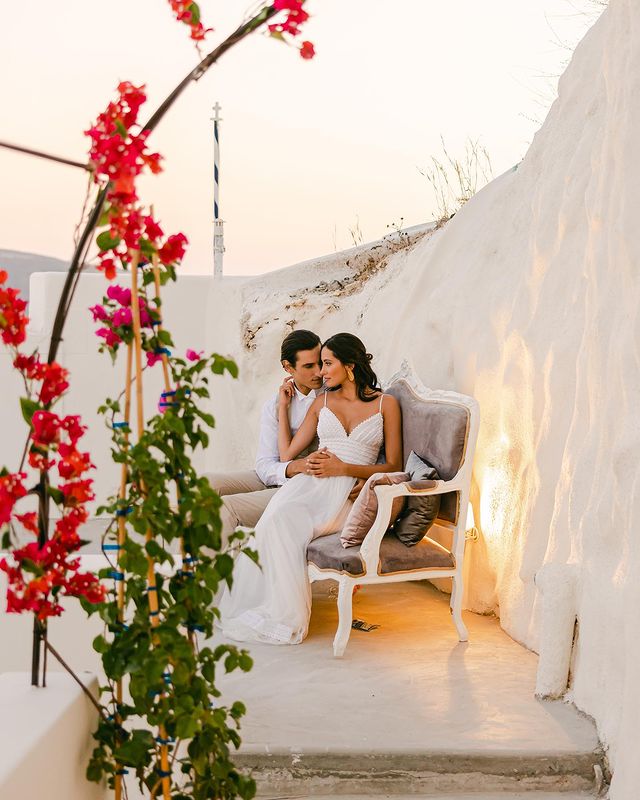 Couple posing in Cycladic backdrop during their destination wedding, framed by red bougainvillea, with bride dressed in a minimal Zolotas gown